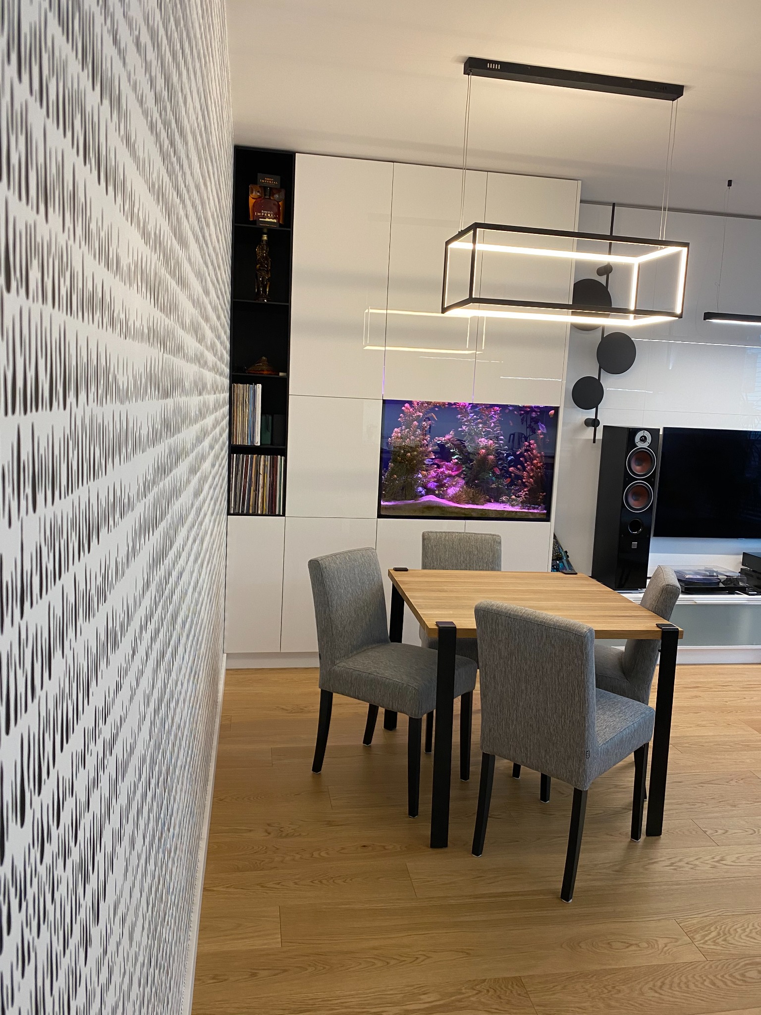 Nowoczesna jadalnia z drewnianym stołem, szarymi krzesłami i akwarium w zabudowie meblowej, oświetlona geometryczną lampą wiszącą.