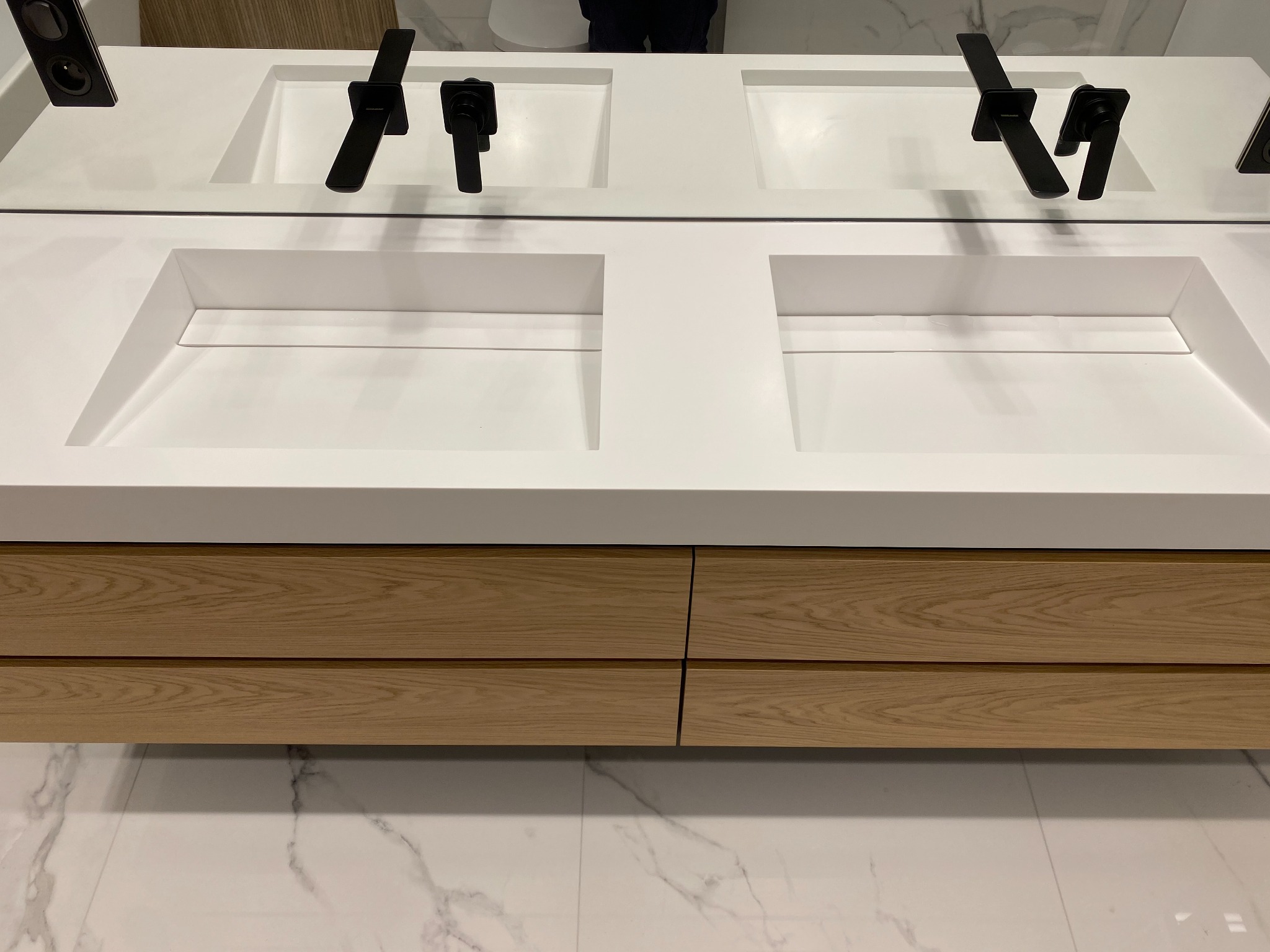 Nowoczesna łazienka z podwójną, wpuszczaną umywalką z białego konglomeratu, czarną armaturą i szafką z jasnego drewna na tle marmurowej podłogi.