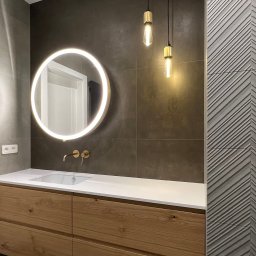 Maria Green Design - Nowoczesna łazienka z drewnianą szafką pod umywalką, okrągłym lustrem z podświetleniem LED i dwoma wiszącymi lampami w stylu retro na tle grafitowych płytek.