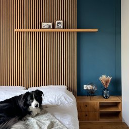 Maria Green Design - Sypialnia z drewnianą ścianą z listew, półką z ramkami, nocnym stolikiem z lampką i wazonem, oraz psem rasy border collie leżącym na łóżku.