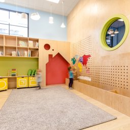 Maria Green Design - Jasne wnętrze pokoju zabaw z drewnianymi meblami, regałami z książkami, kolorową mapą świata na ścianie i bawiącymi się dziećmi w tle.