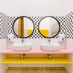 Nowoczesna łazienka z dwoma umywalkami na różowym blacie, żółte półki i wieszaki, ściany w biało-czarne kropki, okrągłe lustra.