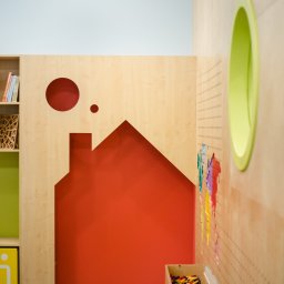 Maria Green Design - Pokój dziecięcy z drewnianymi elementami w kształcie domu, koła i owalu, pomalowany na czerwono, zielono i żółto, z półkami na książki i pojemnikami na zabawki.