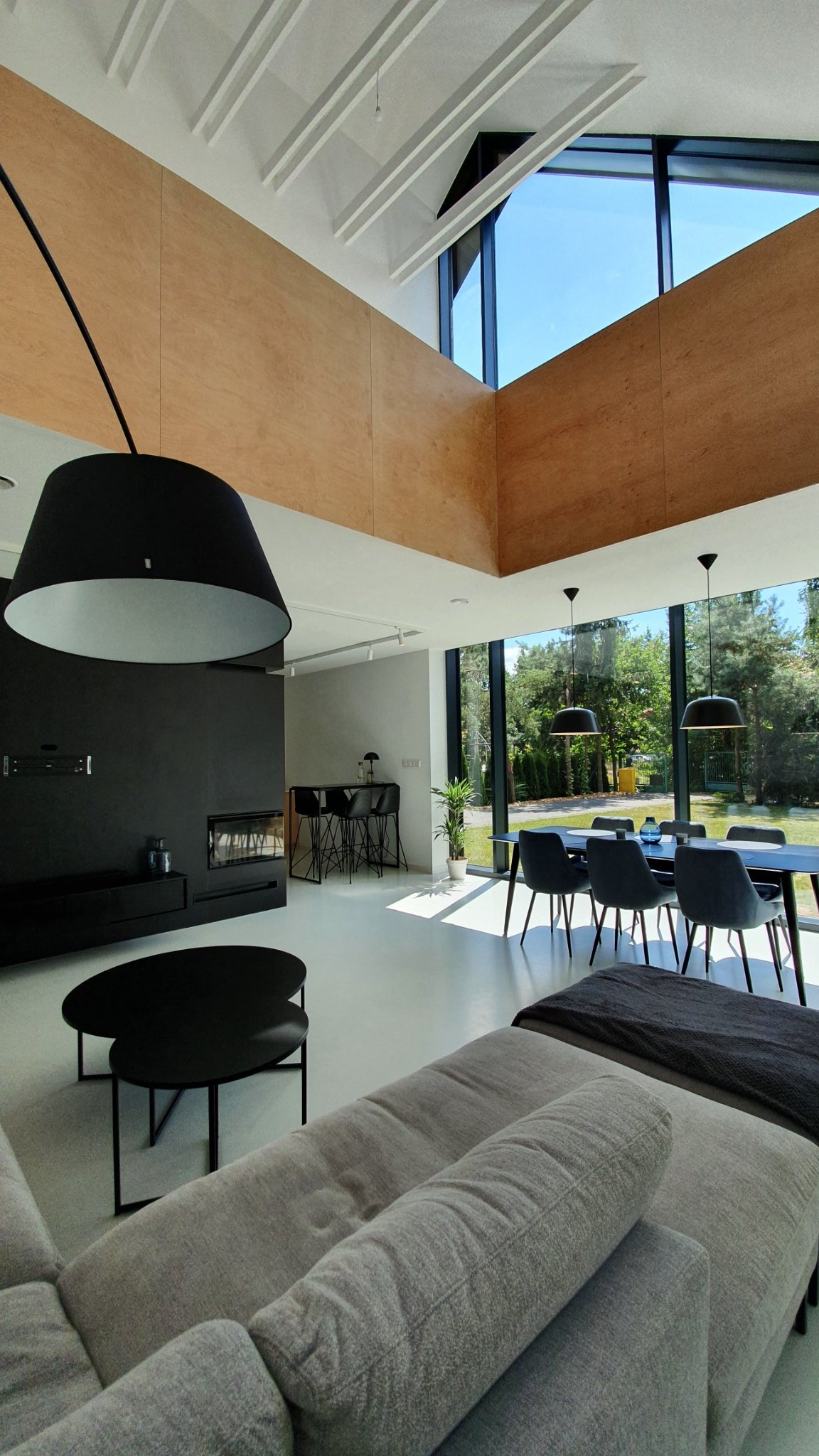 Jasne, przestronne wnętrze domu z wysokim sufitem, drewnianymi panelami i dużymi oknami wychodzącymi na ogród. Wnętrze urządzone w minimalistycznym stylu z czarnymi akcentami, w tym lampami...