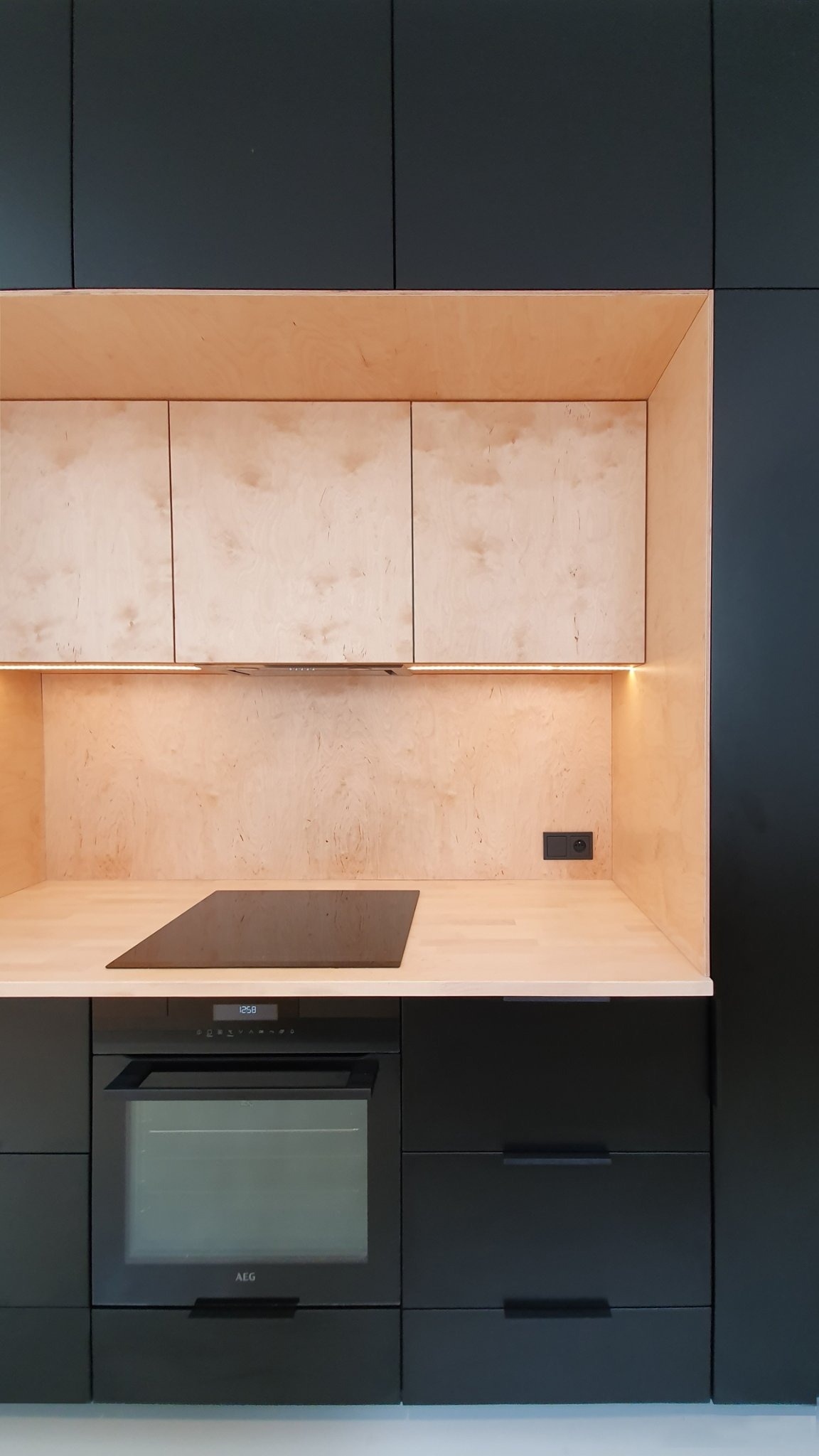 Nowoczesna kuchnia z czarnymi szafkami i elementami z jasnej sklejki, podświetlenie LED pod górnymi szafkami, płyta indukcyjna i piekarnik AEG.
