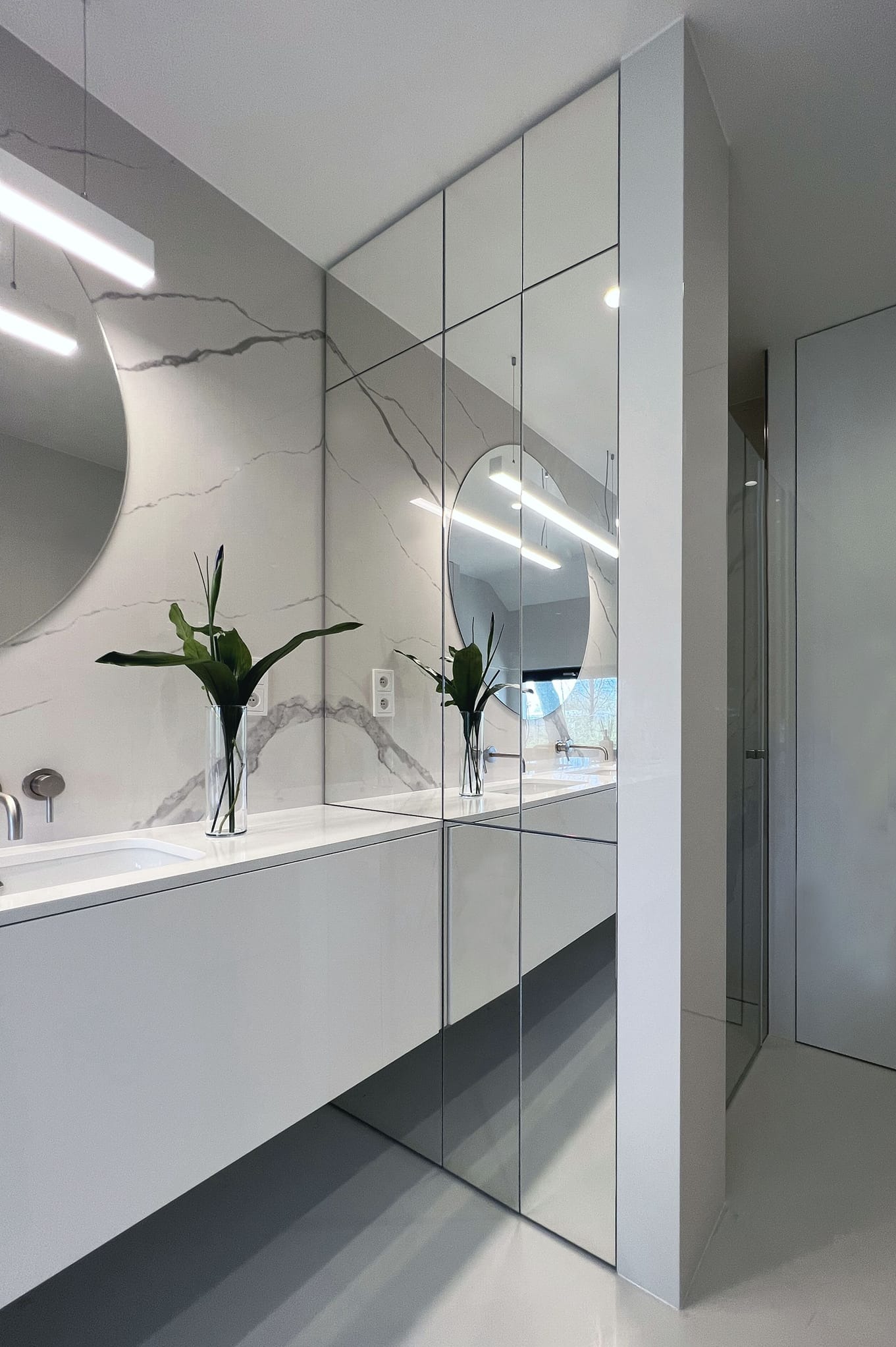 Nowoczesna, minimalistyczna łazienka z białymi szafkami pod umywalką, okrągłym lustrem z podświetleniem LED, ścianą wykończoną płytkami imitującymi marmur z szarymi żyłami oraz dekoracyjnym wazonem...