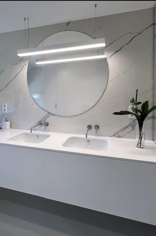 Minimalistyczna łazienka z podwójną umywalką, okrągłym lustrem podświetlonym dwoma poziomymi lampami LED i marmurową ścianą w tle. W wazonie stoi zielona roślina.