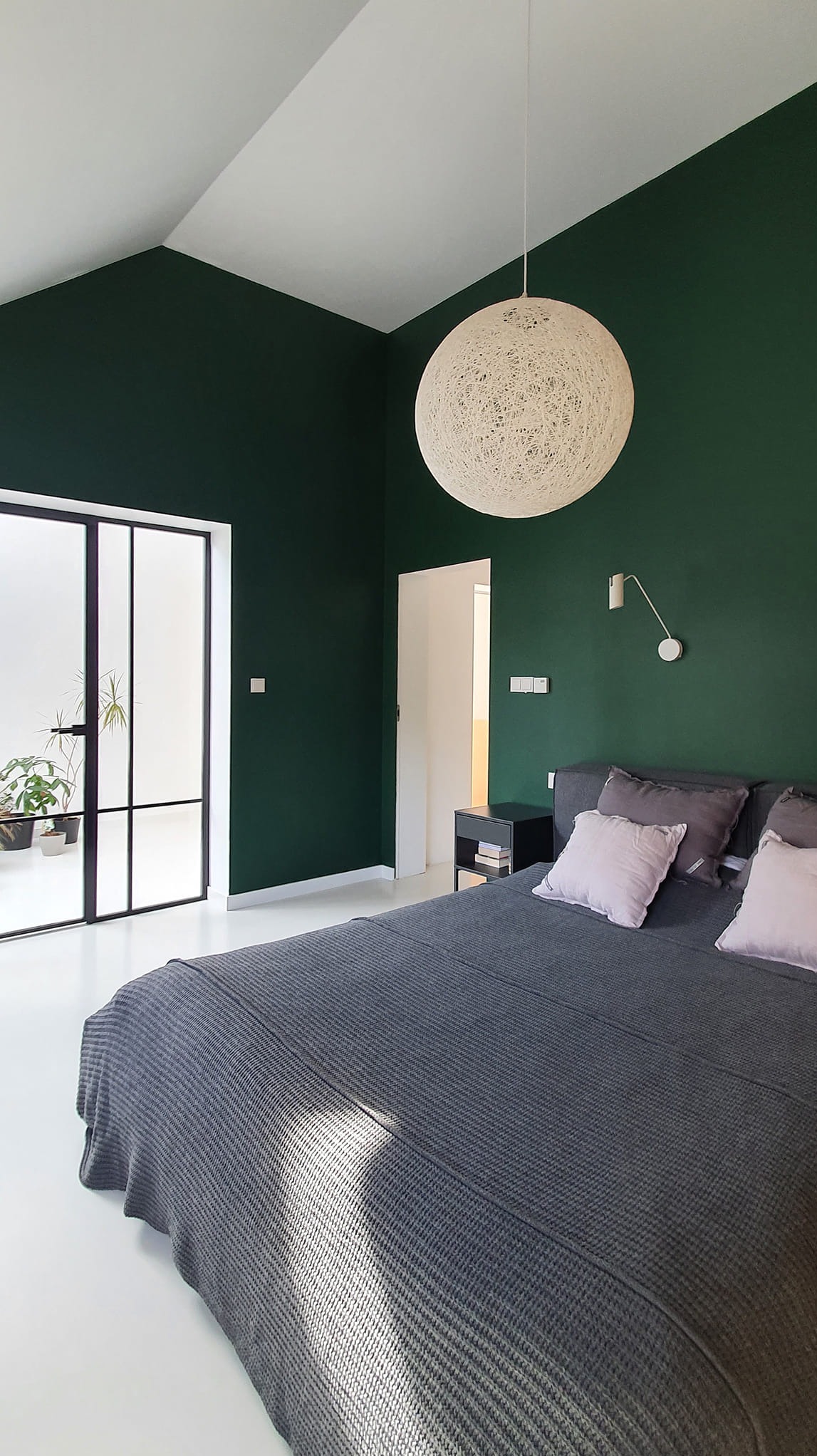 Nowoczesna sypialnia z ciemnozielonymi ścianami, białymi podłogami i dużym, okrągłym abażurem z włókna, minimalistyczna lampka ścienna i widok na patio przez przeszklone drzwi.