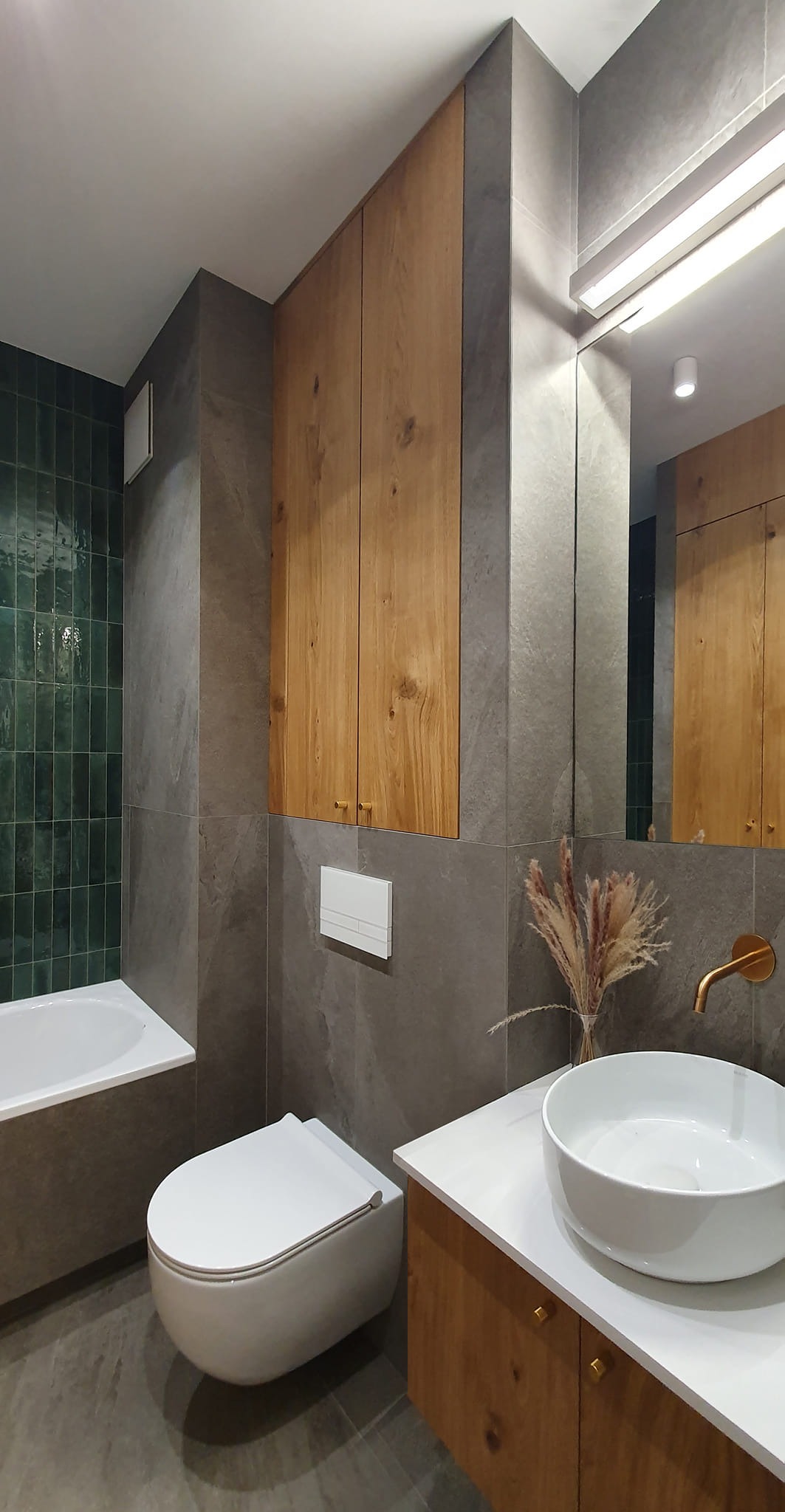 Nowoczesna łazienka z zielonymi płytkami w strefie prysznica, szarymi płytkami na ścianach, drewnianymi szafkami wiszącymi i pod umywalką, białą ceramiką i złotą armaturą.