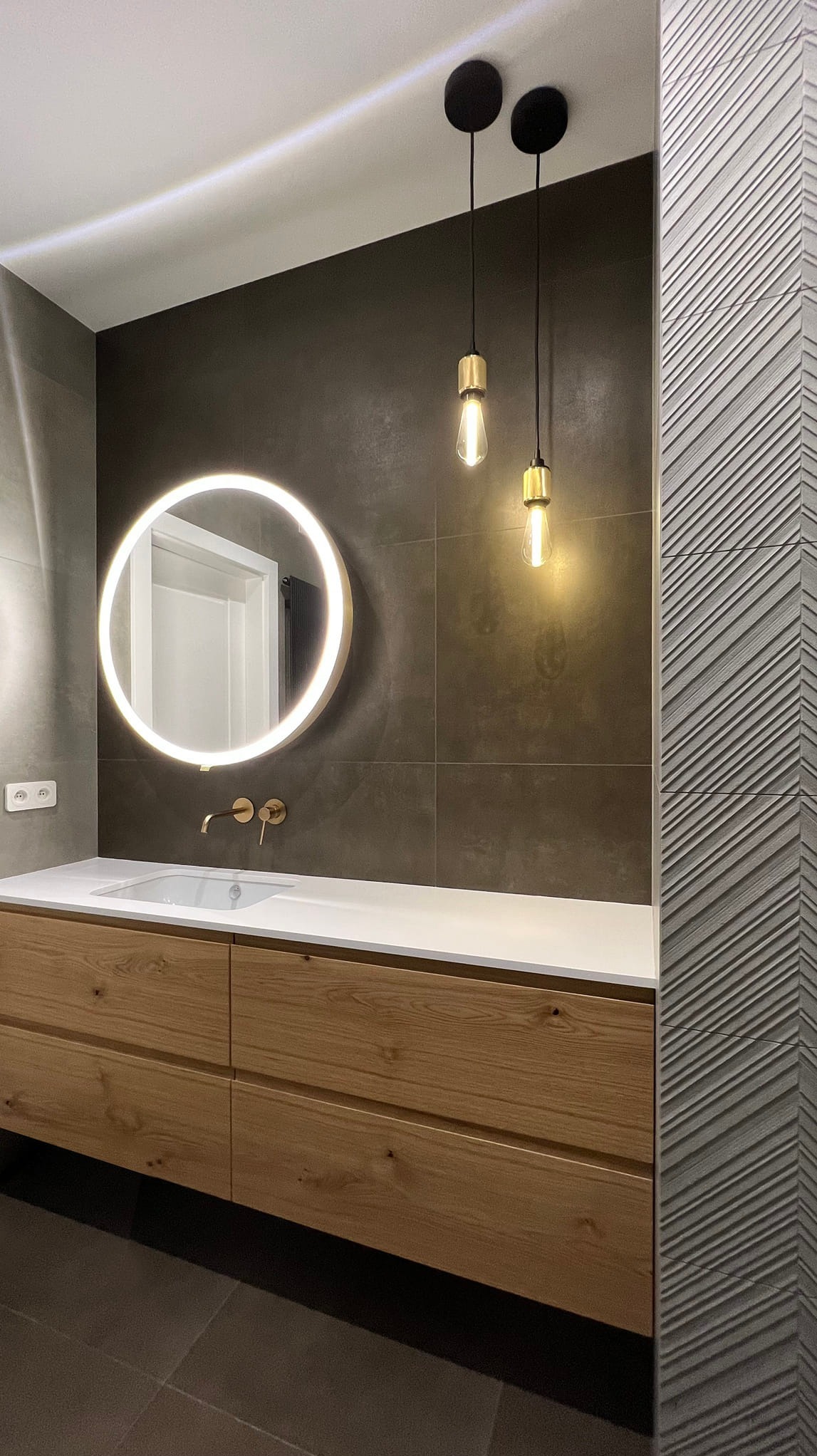 Nowoczesna łazienka z drewnianą szafką pod umywalką, okrągłym lustrem z podświetleniem LED i dwoma wiszącymi lampami w stylu retro na tle grafitowych płytek.