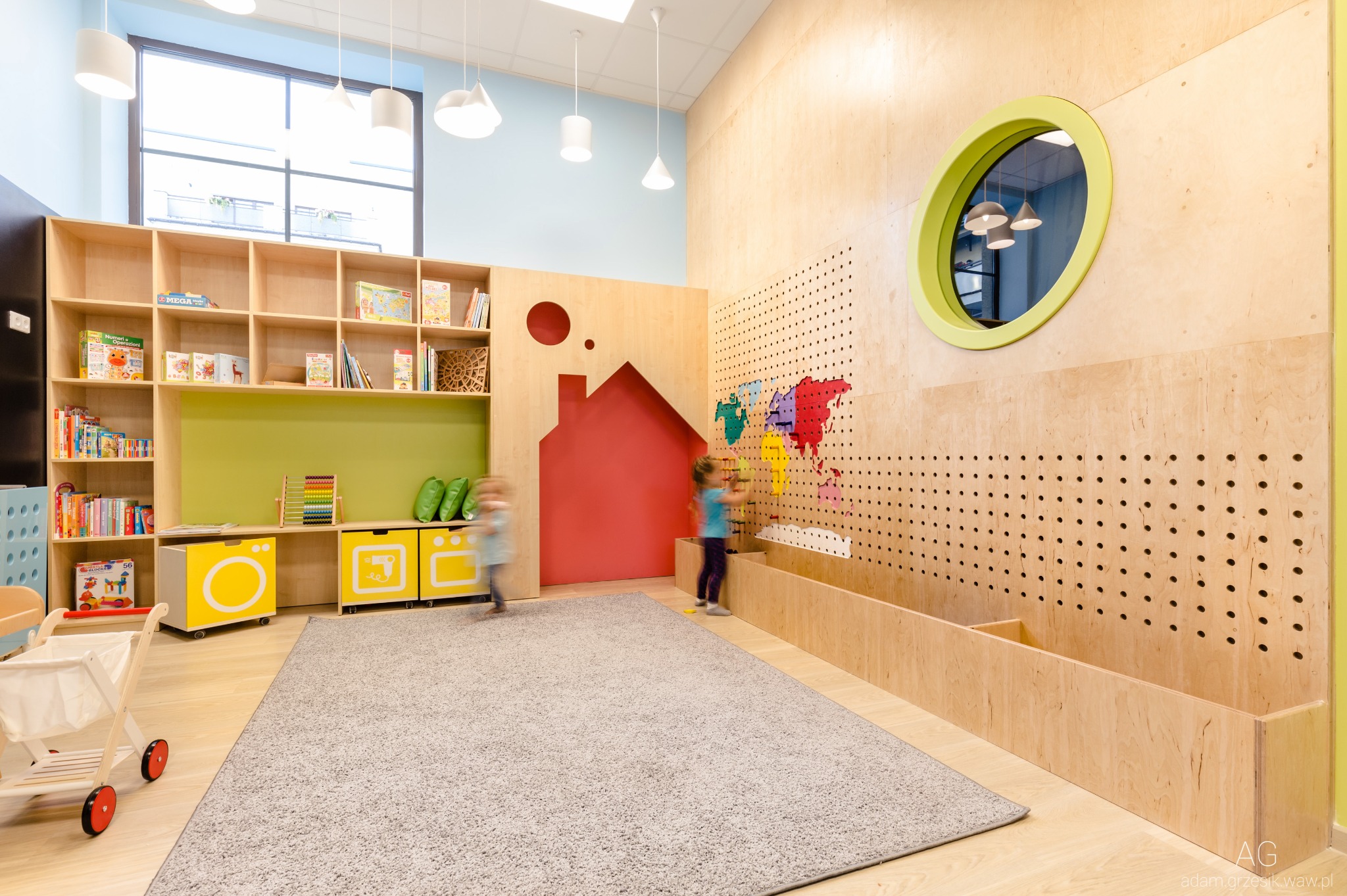 Jasne wnętrze pokoju zabaw z drewnianymi meblami, regałami z książkami, kolorową mapą świata na ścianie i bawiącymi się dziećmi w tle.