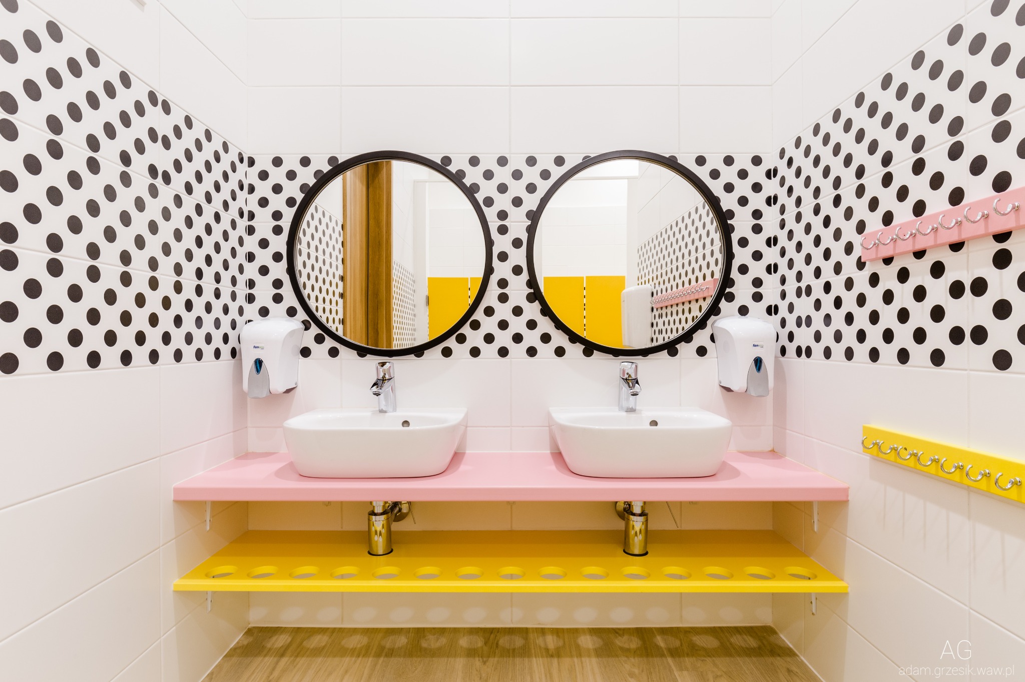 Nowoczesna łazienka z dwoma umywalkami na różowym blacie, żółte półki i wieszaki, ściany w biało-czarne kropki, okrągłe lustra.