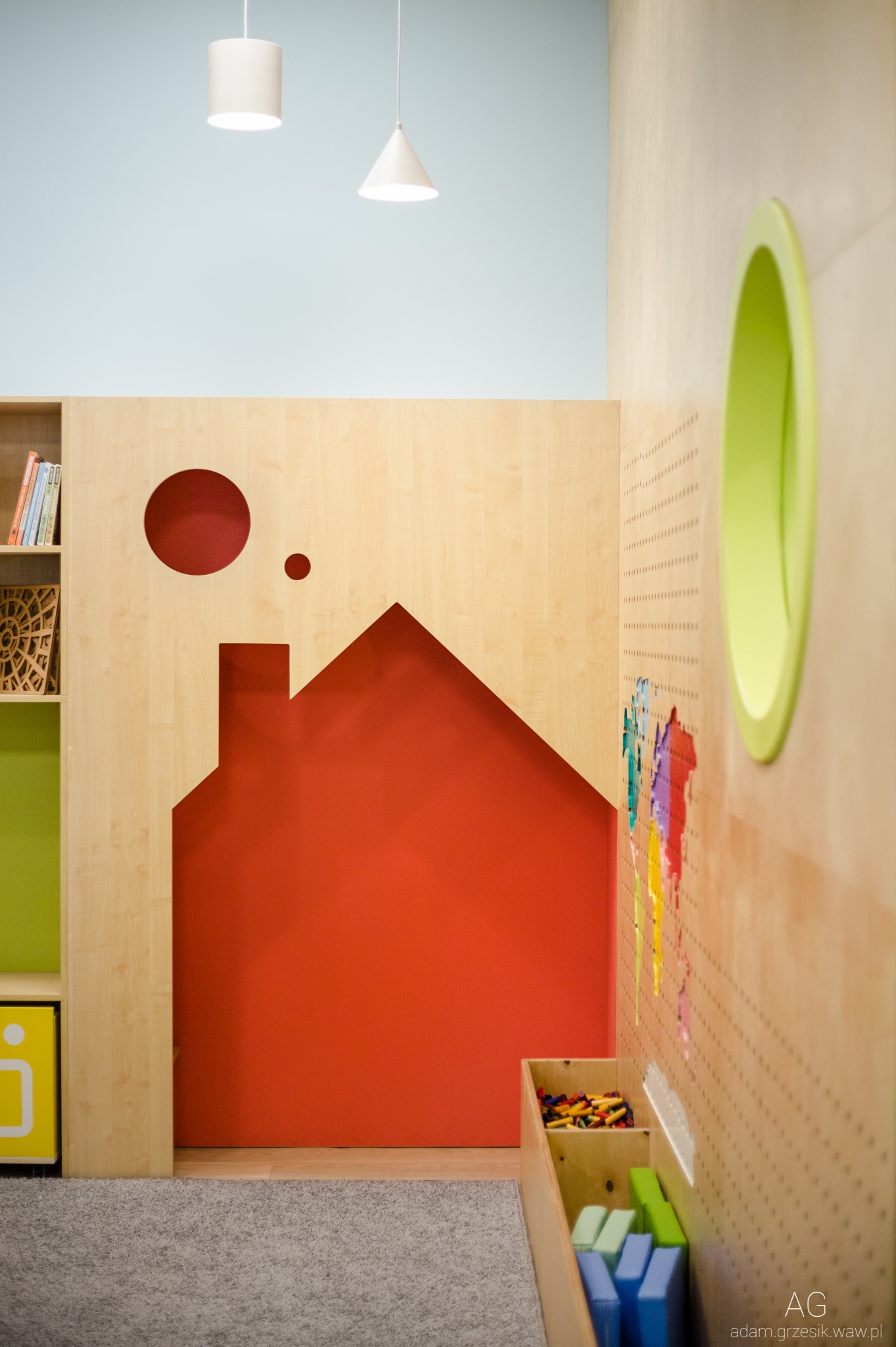 Pokój dziecięcy z drewnianymi elementami w kształcie domu, koła i owalu, pomalowany na czerwono, zielono i żółto, z półkami na książki i pojemnikami na zabawki.