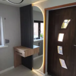 Nowoczesny przedpokój z drewnianą konsolą z szufladą, lamelami i podświetlanym lustrem w kształcie łuku, obok brązowe drzwi wejściowe z geometrycznymi szybkami.