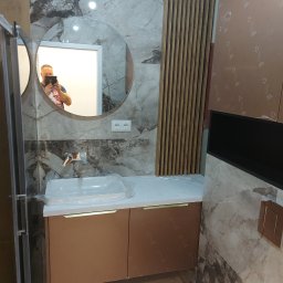 TS STYL MEBLE TOMASZ SIWCZYK - Nowoczesna łazienka w trakcie wykańczania: marmurowe płytki, okrągłe lustro z odbiciem fotografa, umywalka z szafką w kolorze miedzi, drewniane lamele dekoracyjne na ścianie.