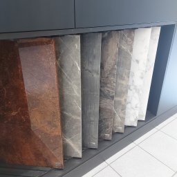 TS STYL MEBLE TOMASZ SIWCZYK - Ekspozycja próbek blatów kuchennych z różnymi wzorami marmuru i drewna, umieszczonych w szufladzie pod szafką kuchenną.