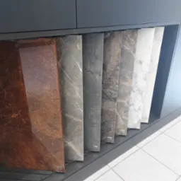 Ekspozycja próbek blatów kuchennych z różnymi wzorami marmuru i drewna, umieszczonych w szufladzie pod szafką kuchenną.