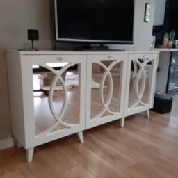 Biała komoda RTV z lustrzanymi frontami i dekoracyjnymi, okrągłymi wycięciami, stojąca na drewnianej podłodze, z telewizorem na blacie.