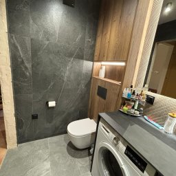 TS STYL MEBLE TOMASZ SIWCZYK - Nowoczesna łazienka z szarymi płytkami imitującymi marmur, podwieszaną toaletą, zabudową z drewnianymi szafkami z oświetleniem LED i pralką pod blatem.