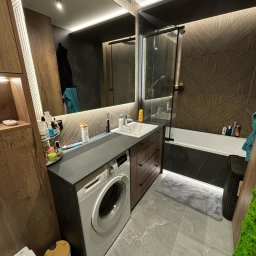 TS STYL MEBLE TOMASZ SIWCZYK - Nowoczesna łazienka z pralką zabudowaną pod blatem, drewniane szafki z szufladami, duża wanna za szklaną ścianką, wzorzyste brązowe płytki na ścianie, szara podłoga i oświetlenie LED.