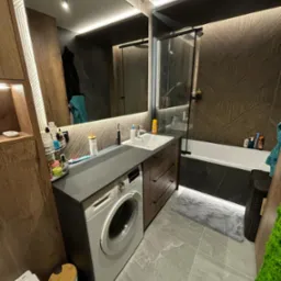 Nowoczesna łazienka z pralką zabudowaną pod blatem, drewniane szafki z szufladami, duża wanna za szklaną ścianką, wzorzyste brązowe płytki na ścianie, szara podłoga i oświetlenie LED.