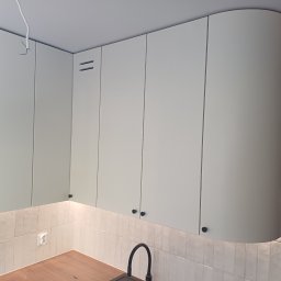 TS STYL MEBLE TOMASZ SIWCZYK - Jasne, minimalistyczne szafki kuchenne podwieszane z zaokrąglonym narożnikiem, czarne gałki, płytki imitujące cegłę na ścianie, widoczny fragment blatu i zlew. W tle widoczny fragment osoby.