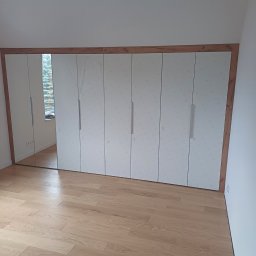 TS STYL MEBLE TOMASZ SIWCZYK - Biała szafa wnękowa z lustrzanymi drzwiami, obramowana drewnem, na podłodze z jasnego drewna, częściowo jeszcze w folii ochronnej.