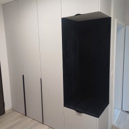 TS STYL MEBLE TOMASZ SIWCZYK - Szara szafa na wymiar z wnęką do siedzenia wyłożoną czarnym materiałem. Minimalistyczny design, jasne wnętrze, widoczny fragment drzwi.