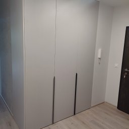 TS STYL MEBLE TOMASZ SIWCZYK - Jasnoszara szafa wnękowa z trzema drzwiami i minimalistycznymi uchwytami, dopasowana do jasnego wnętrza z parkietem i drzwiami po prawej stronie.