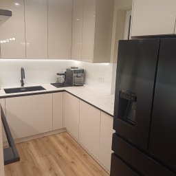 TS STYL MEBLE TOMASZ SIWCZYK - Jasna kuchnia z połyskiem: białe szafki, czarny zlew i armatura, marmurowy blat, ekspres do kawy i czarna lodówka side-by-side. Drewniana podłoga.