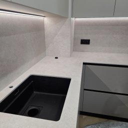 TS STYL MEBLE TOMASZ SIWCZYK - Minimalistyczna kuchnia z jasnoszarym, nakrapianym blatem i panelem ściennym, zintegrowanym czarnym zlewem oraz subtelnym oświetleniem LED pod szafkami. Widoczne czarne gniazdka i wysuwane gniazdo...