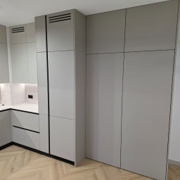 TS STYL MEBLE TOMASZ SIWCZYK - Jasnoszara zabudowa meblowa w kuchni i przedpokoju, z bezuchwytowymi frontami i czarnymi liniami podziału, na tle podłogi w jodełkę. Widoczne oświetlenie podszafkowe i kratki wentylacyjne.