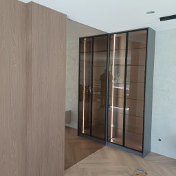 TS STYL MEBLE TOMASZ SIWCZYK - Nowoczesne witryny z przyciemnianymi szklanymi drzwiami i wewnętrznym pionowym oświetleniem LED, osadzone w szarych ramach. Obok jasny drewniany panel ścienny, na podłodze parkiet w jodełkę.
