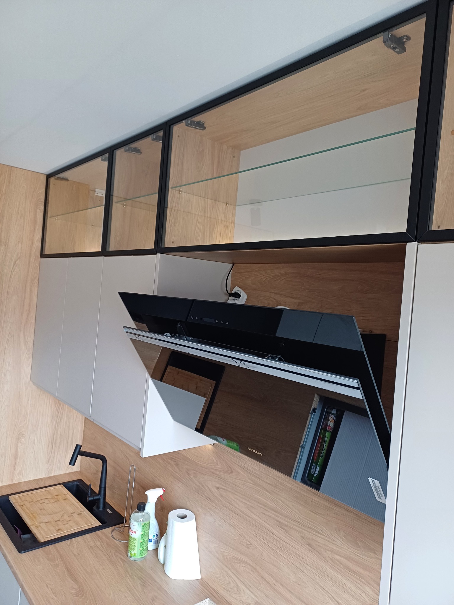 Nowoczesna kuchnia z drewnianym blatem, czarnym zlewem i szklanymi szafkami górnymi z czarnymi ramami, uzupełniona o białe szafki i czarny okap. Na blacie widoczne środki czystości i deska...