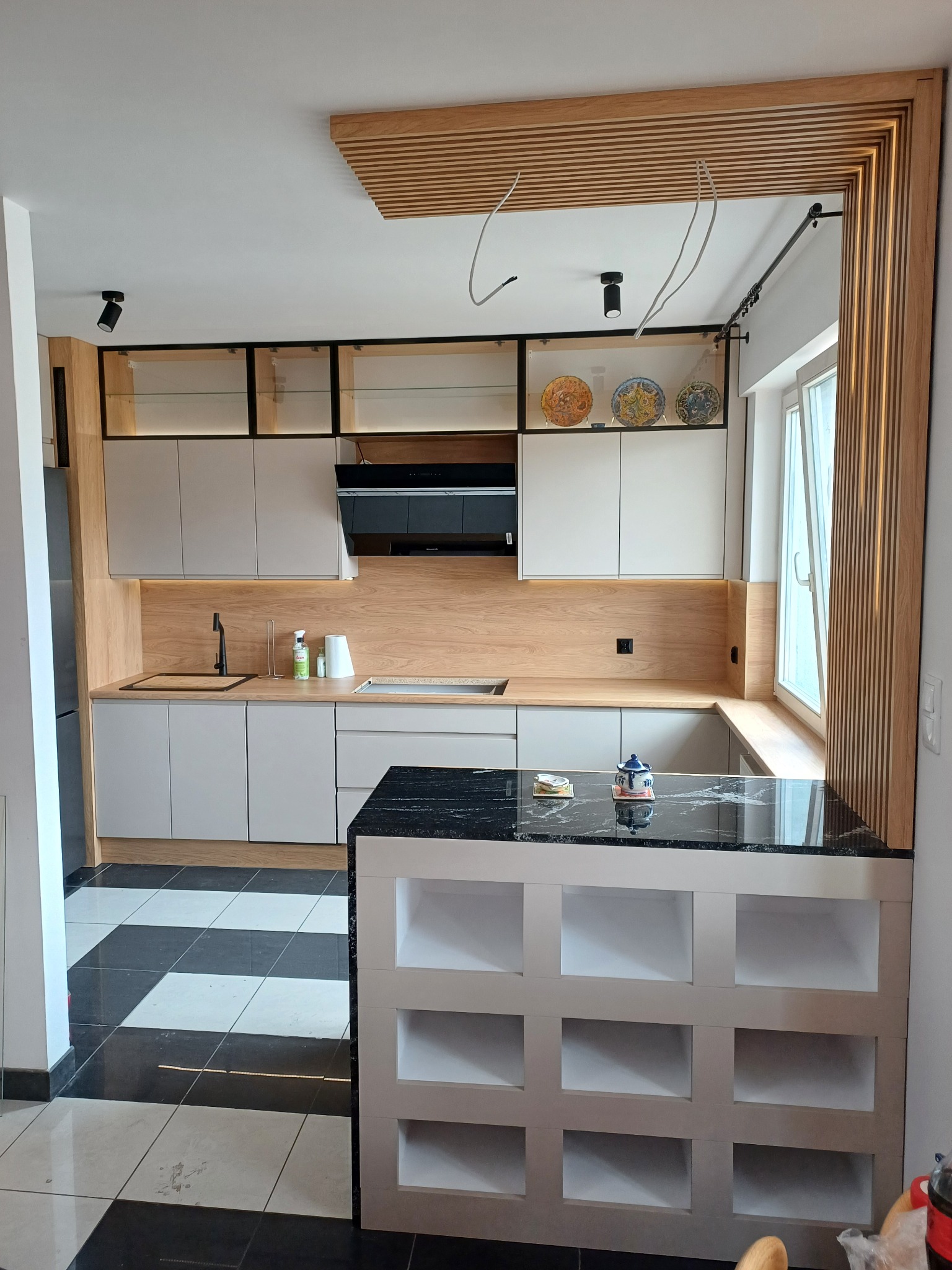 Nowoczesna kuchnia z szarymi szafkami, blatem z czarnego kamienia i drewnianymi elementami dekoracyjnymi, w tym lamele na suficie i ścianie, oraz czarno-białą podłogą w szachownicę.