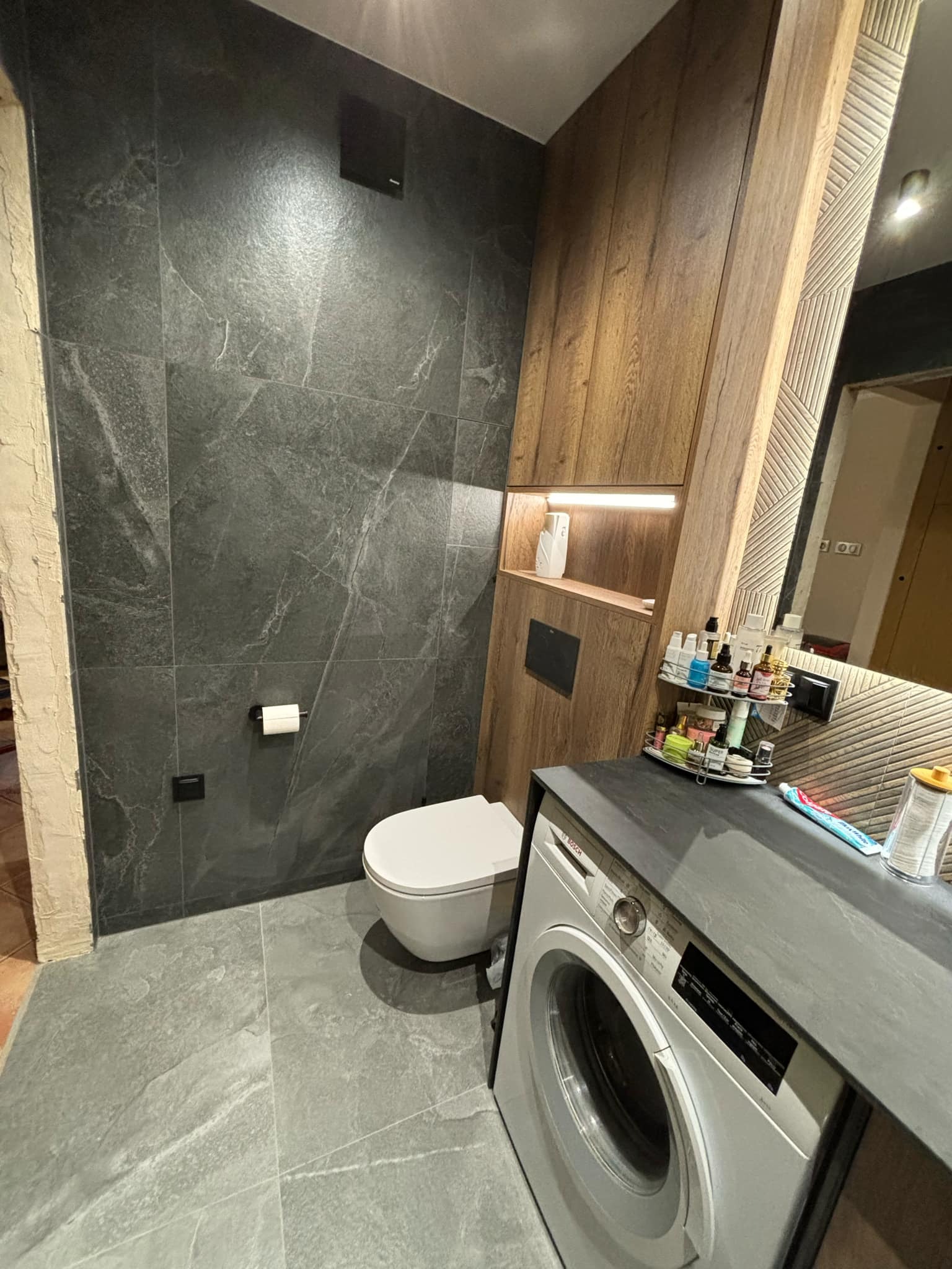 Nowoczesna łazienka z szarymi płytkami imitującymi marmur, podwieszaną toaletą, zabudową z drewnianymi szafkami z oświetleniem LED i pralką pod blatem.