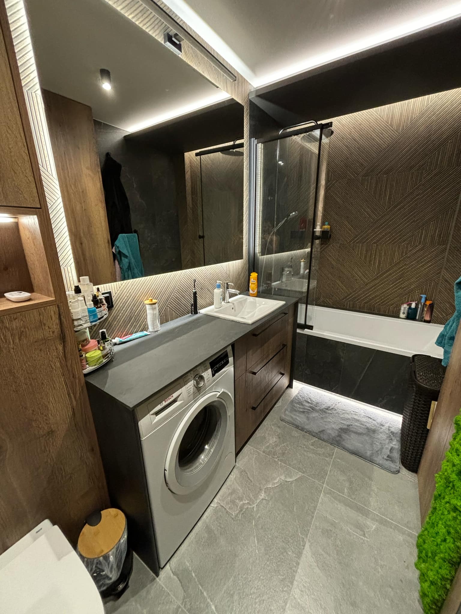 Nowoczesna łazienka z pralką zabudowaną pod blatem, drewniane szafki z szufladami, duża wanna za szklaną ścianką, wzorzyste brązowe płytki na ścianie, szara podłoga i oświetlenie LED.