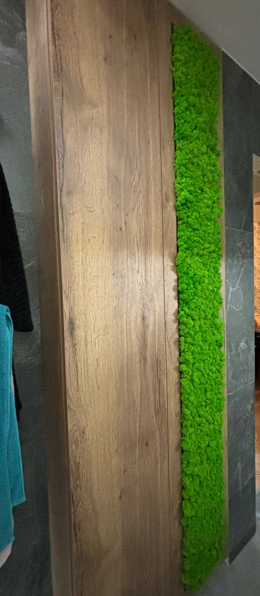 Drewniana szafa na wymiar z pionowym panelem z mchu dekoracyjnego, umieszczona w łazience z szarymi płytkami.