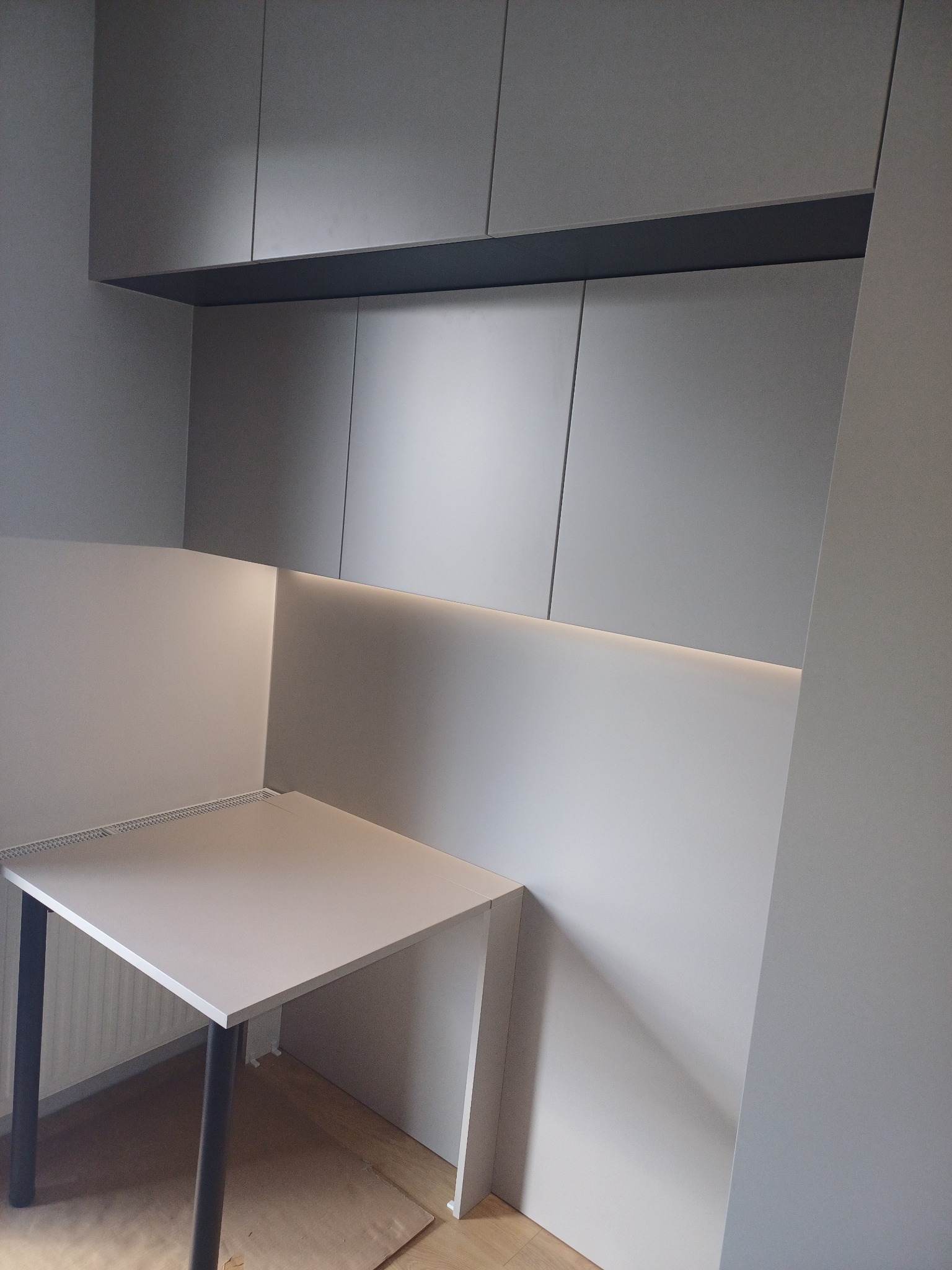 Szare szafki wiszące z podświetleniem LED nad blatem roboczym, minimalistyczny design, widoczne niedoskonałości wykończenia podłogi.