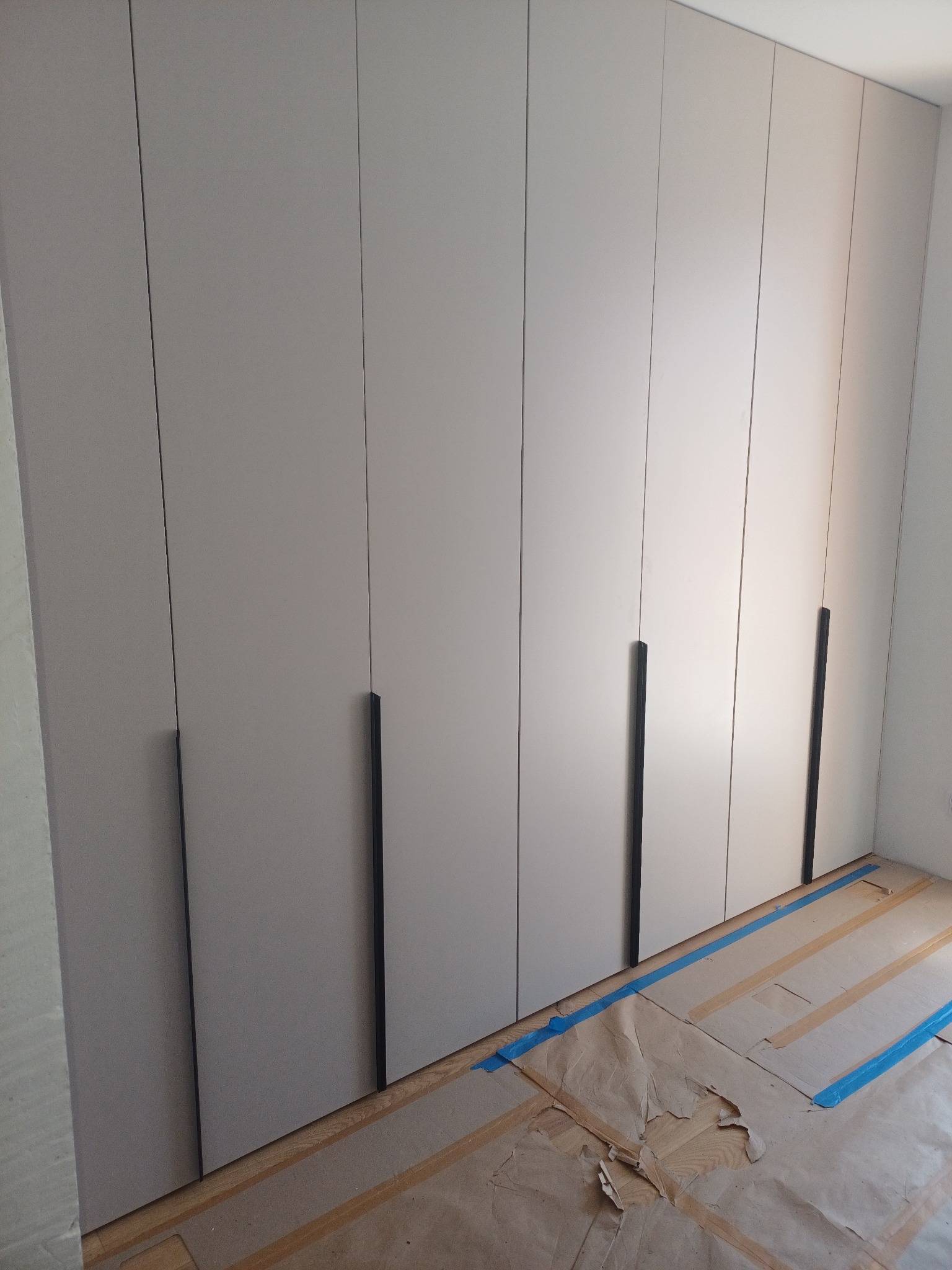Wysoka, szara szafa wnękowa z minimalistycznymi, pionowymi uchwytami, podłoga zabezpieczona kartonem i niebieską taśmą malarską.