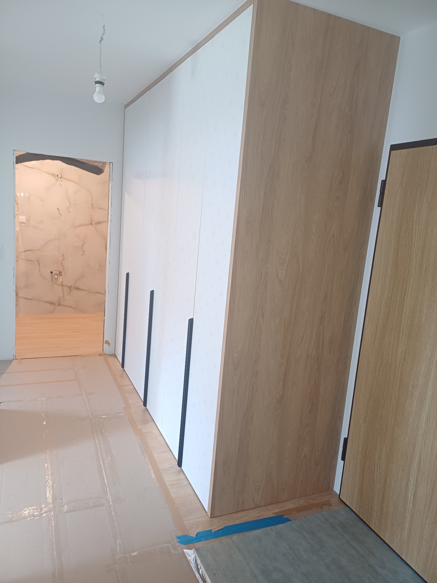 Wnętrze w trakcie wykańczania: biała szafa z ciemnymi, pionowymi uchwytami na tle marmurowej łazienki, podłoga zabezpieczona folią ochronną, widoczna surowa żarówka.