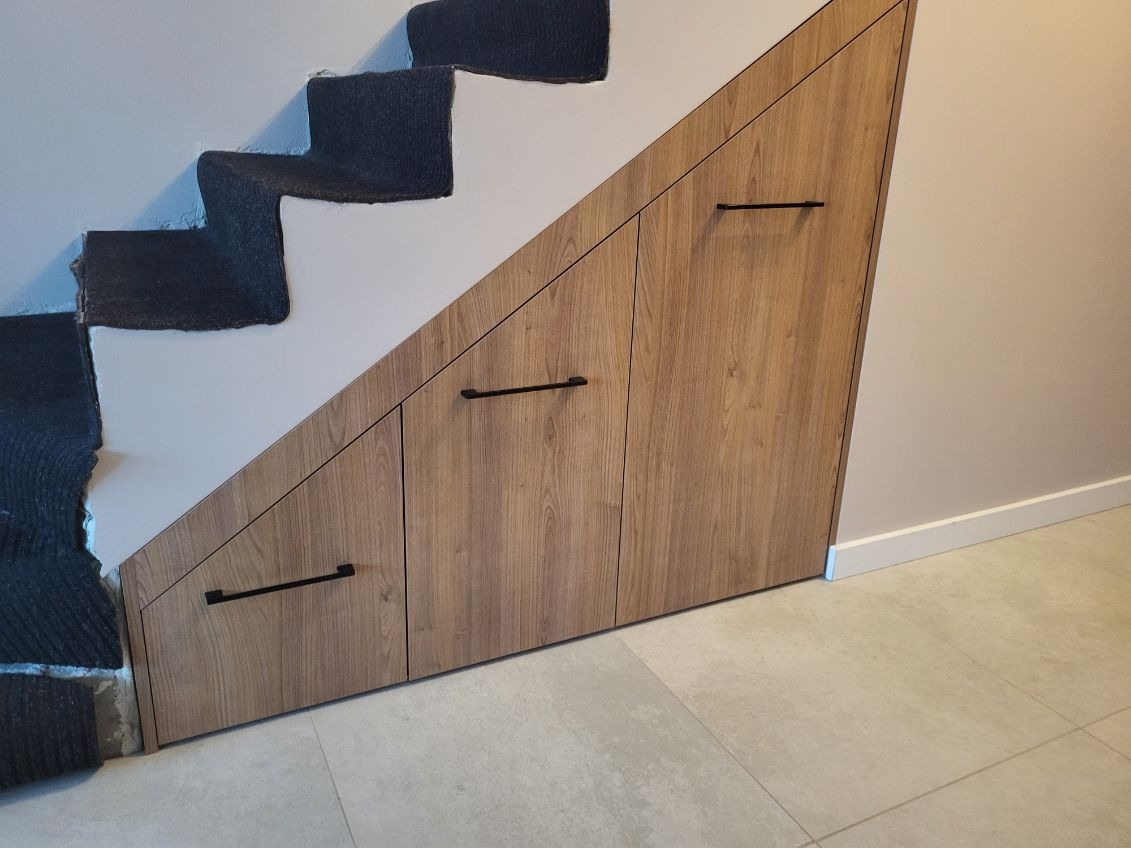Szafka wnękowa pod schodami z drewnopodobnymi frontami i czarnymi uchwytami, widok na fragment schodów z szarym dywanem i jasną podłogą z płytek.
