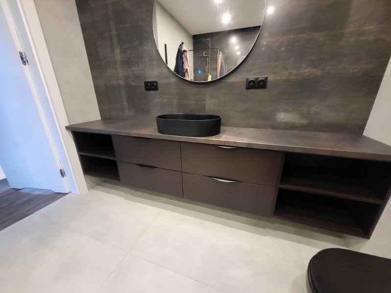 Nowoczesna łazienka z brązowymi szafkami pod umywalką, owalną czarną umywalką nablatową, dużym okrągłym lustrem i ciemną okładziną ściany imitującą kamień.