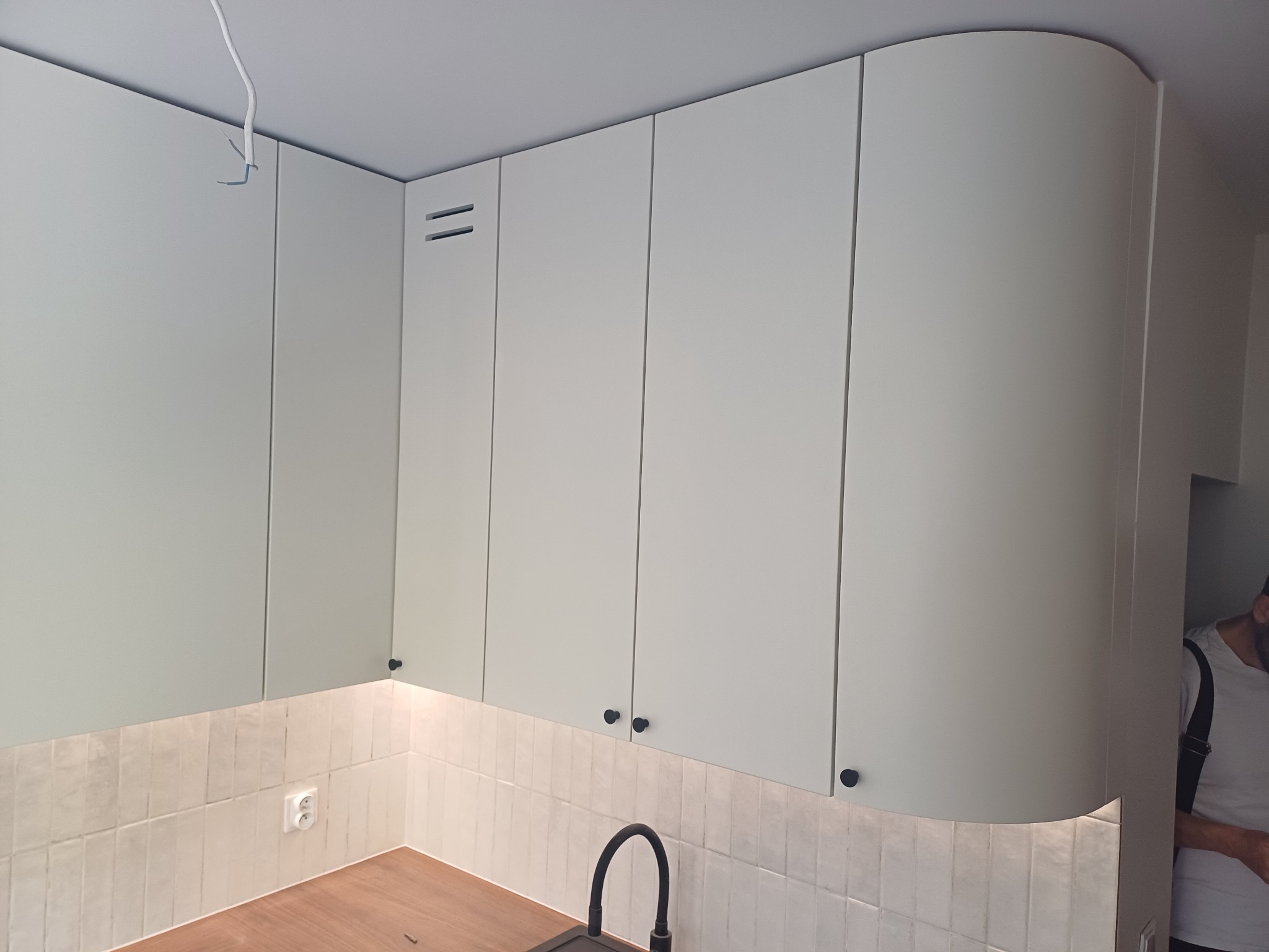 Jasne, minimalistyczne szafki kuchenne podwieszane z zaokrąglonym narożnikiem, czarne gałki, płytki imitujące cegłę na ścianie, widoczny fragment blatu i zlew. W tle widoczny fragment osoby.