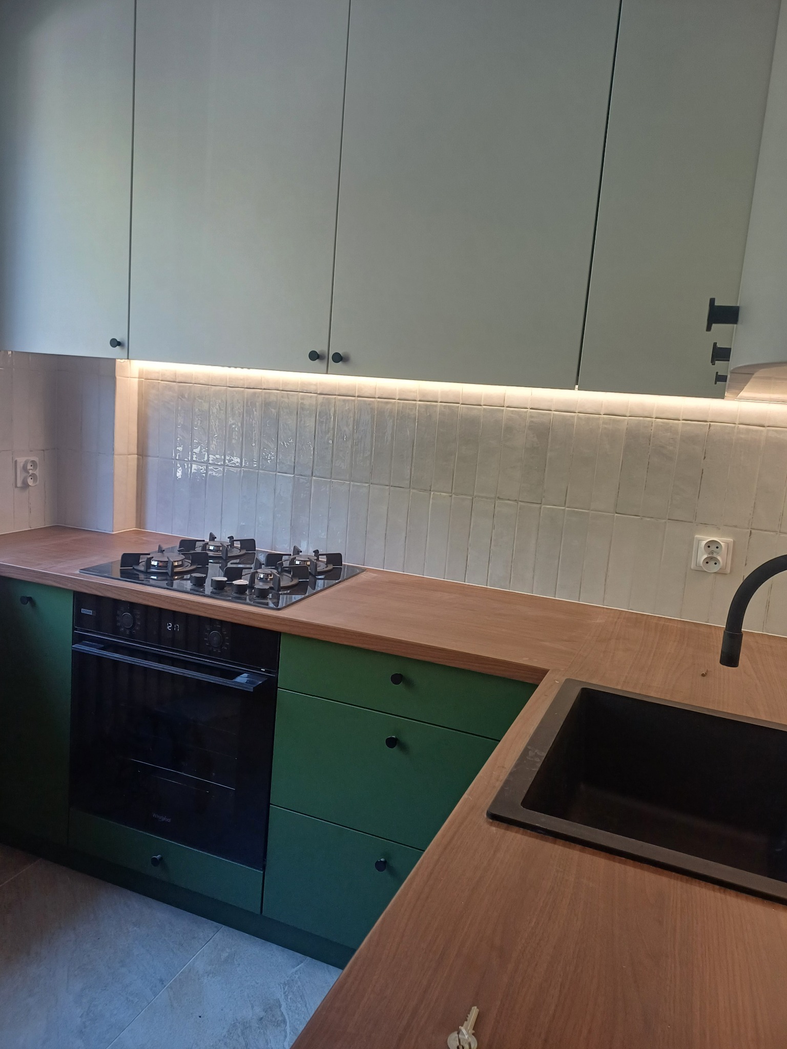 Zielona zabudowa kuchenna z drewnianym blatem, czarnym zlewem i płytą gazową, oświetlenie LED pod szafkami wiszącymi i płytki w stylu cegiełek na ścianie.