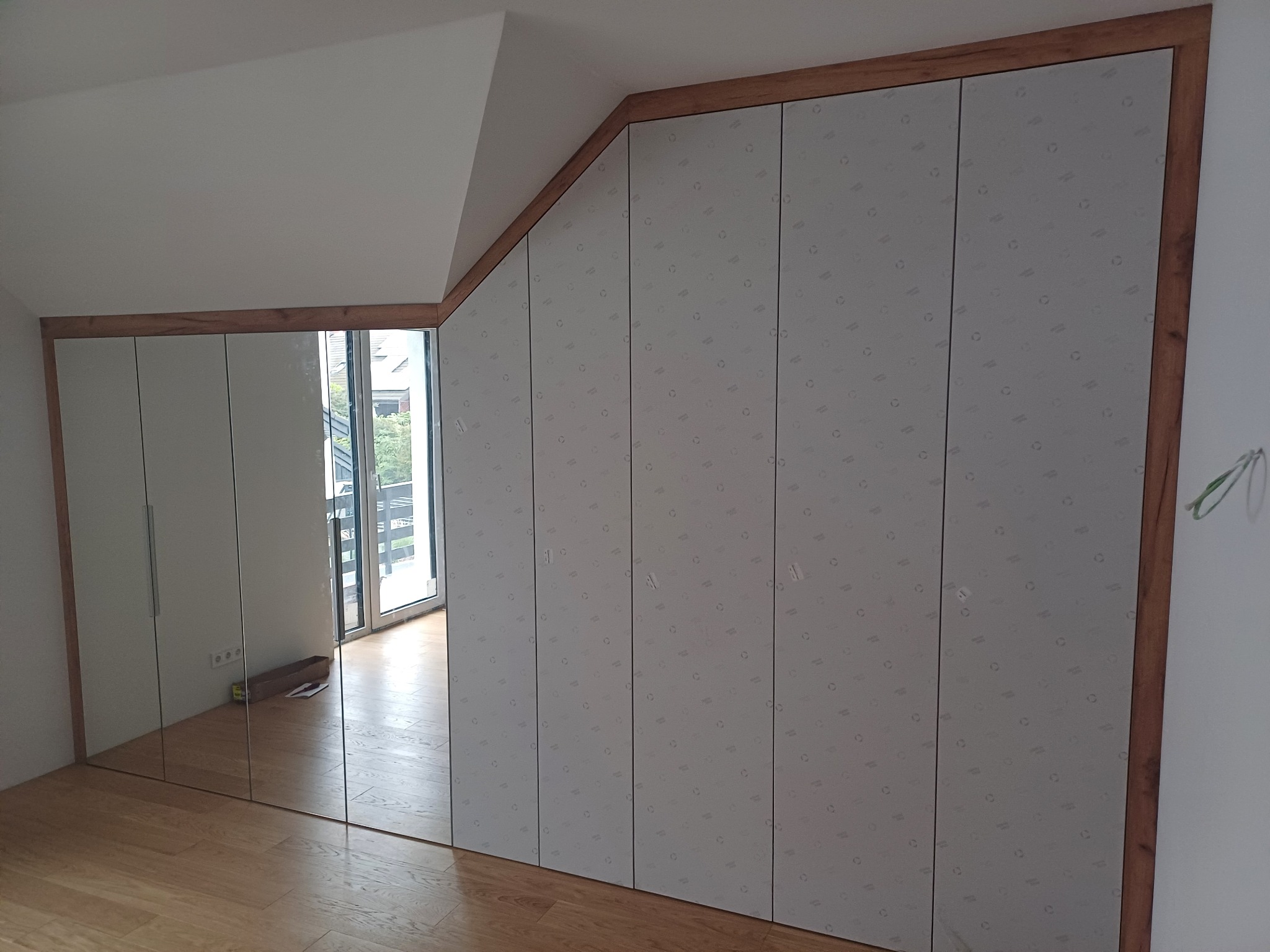 Szafa wnękowa z lustrzanymi drzwiami i drewnianym obramowaniem, dopasowana do skośnego sufitu na poddaszu, z widocznym odbiciem pokoju.