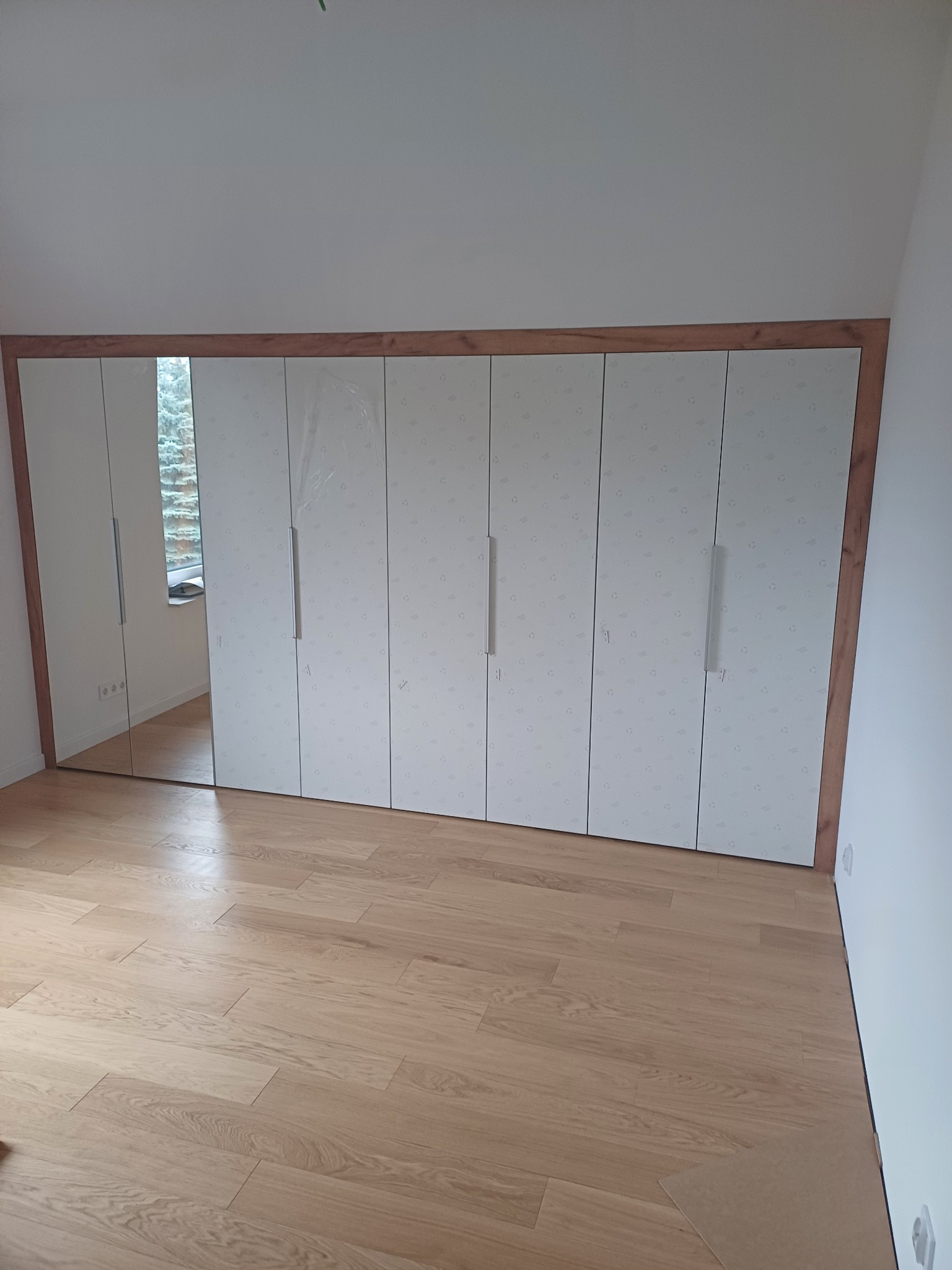 Biała szafa wnękowa z lustrzanymi drzwiami, obramowana drewnem, na podłodze z jasnego drewna, częściowo jeszcze w folii ochronnej.