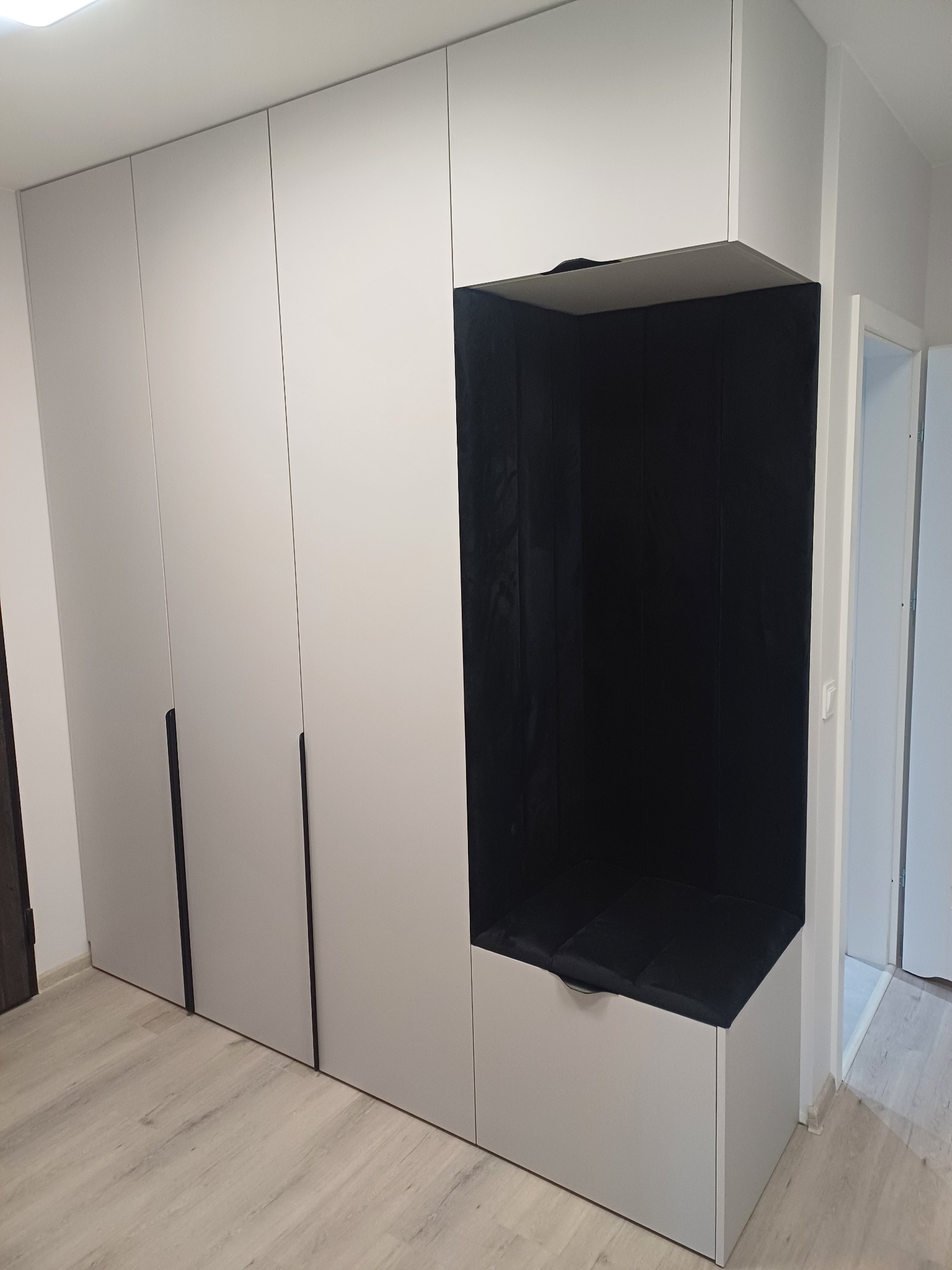 Szara szafa na wymiar z wnęką do siedzenia wyłożoną czarnym materiałem. Minimalistyczny design, jasne wnętrze, widoczny fragment drzwi.