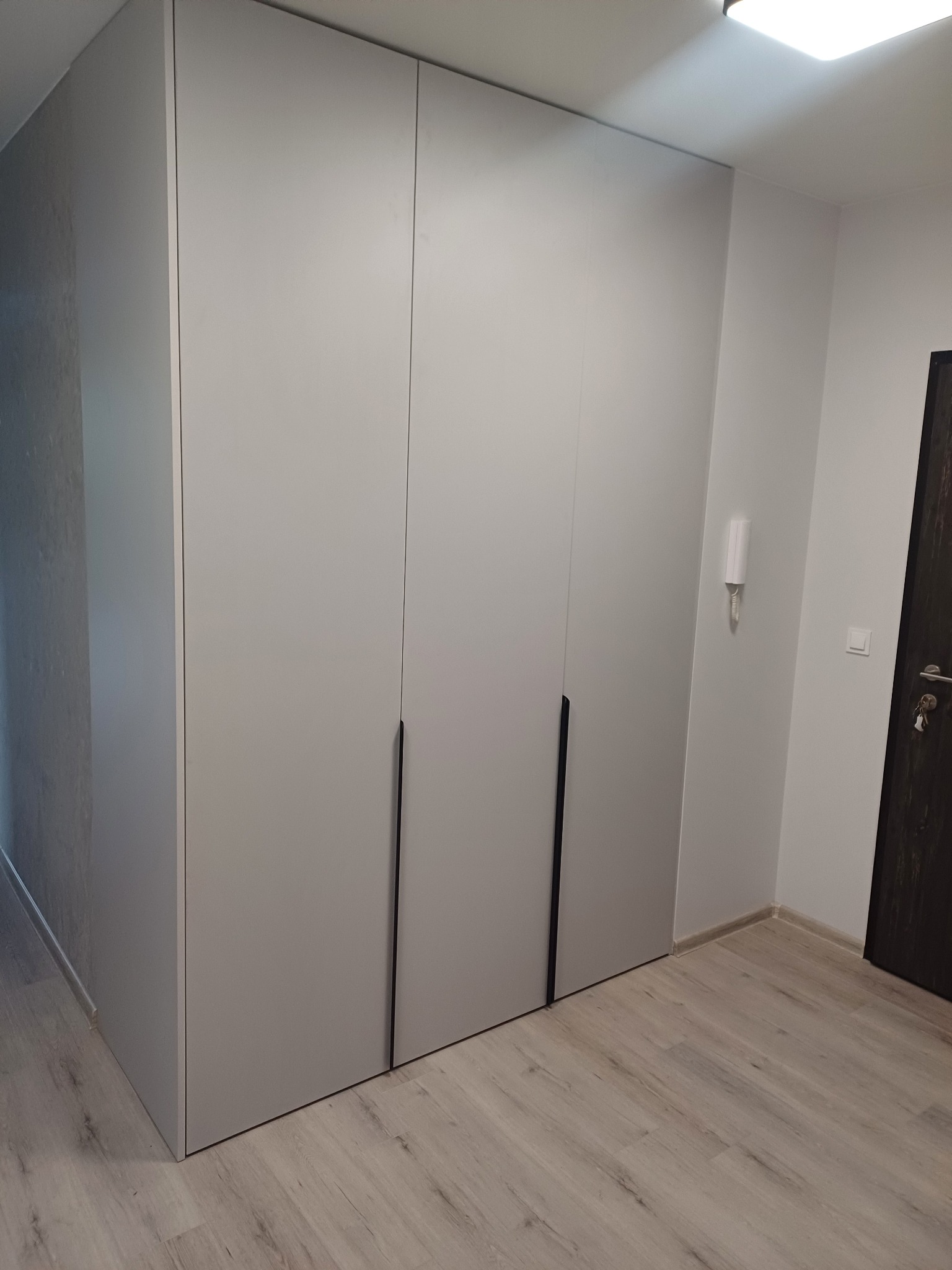 Jasnoszara szafa wnękowa z trzema drzwiami i minimalistycznymi uchwytami, dopasowana do jasnego wnętrza z parkietem i drzwiami po prawej stronie.