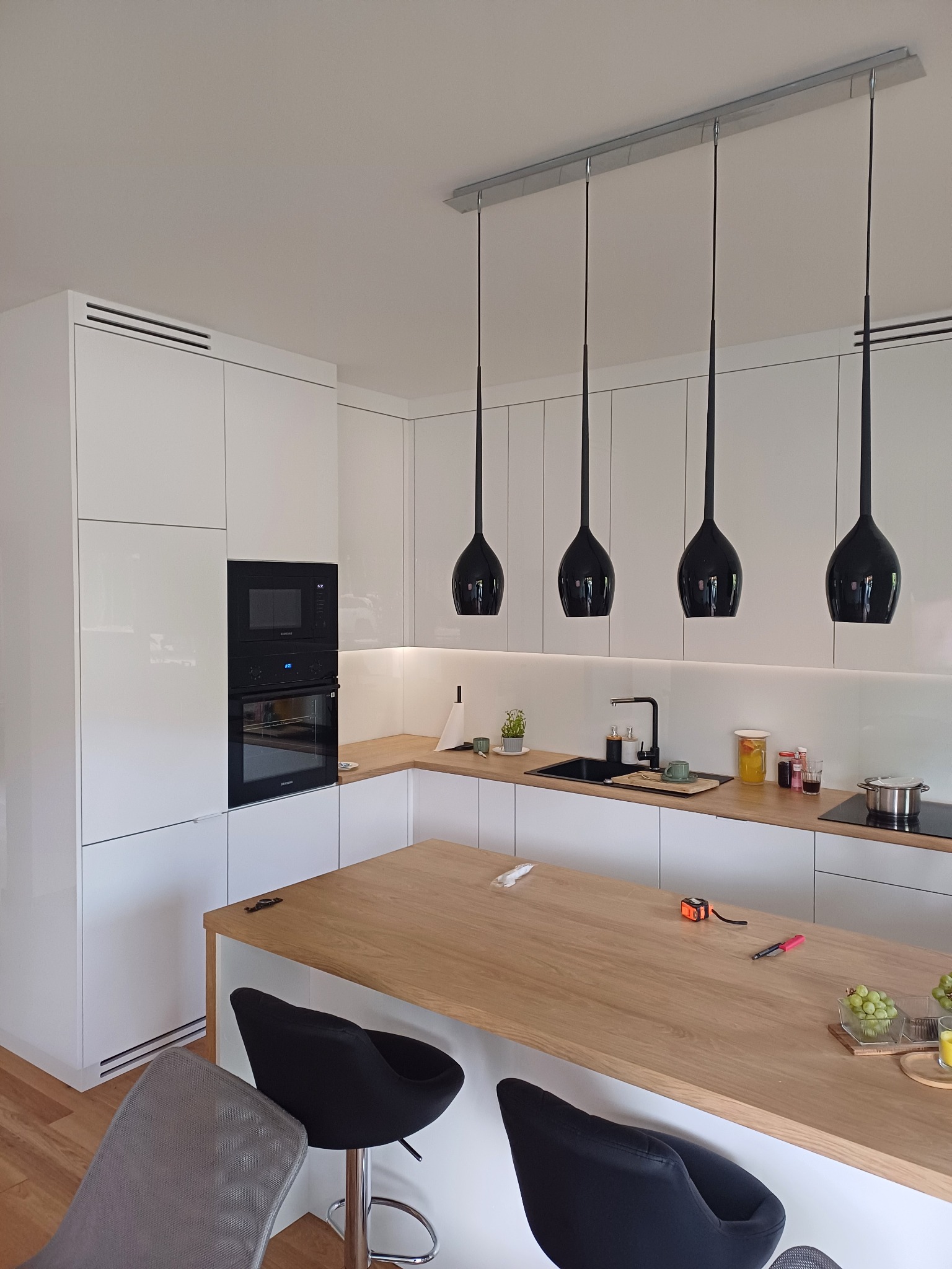 Nowoczesna kuchnia z białymi, lakierowanymi frontami, drewnianym blatem i czarnymi lampami wiszącymi. Na blacie leżą narzędzia pomiarowe i owoce.
